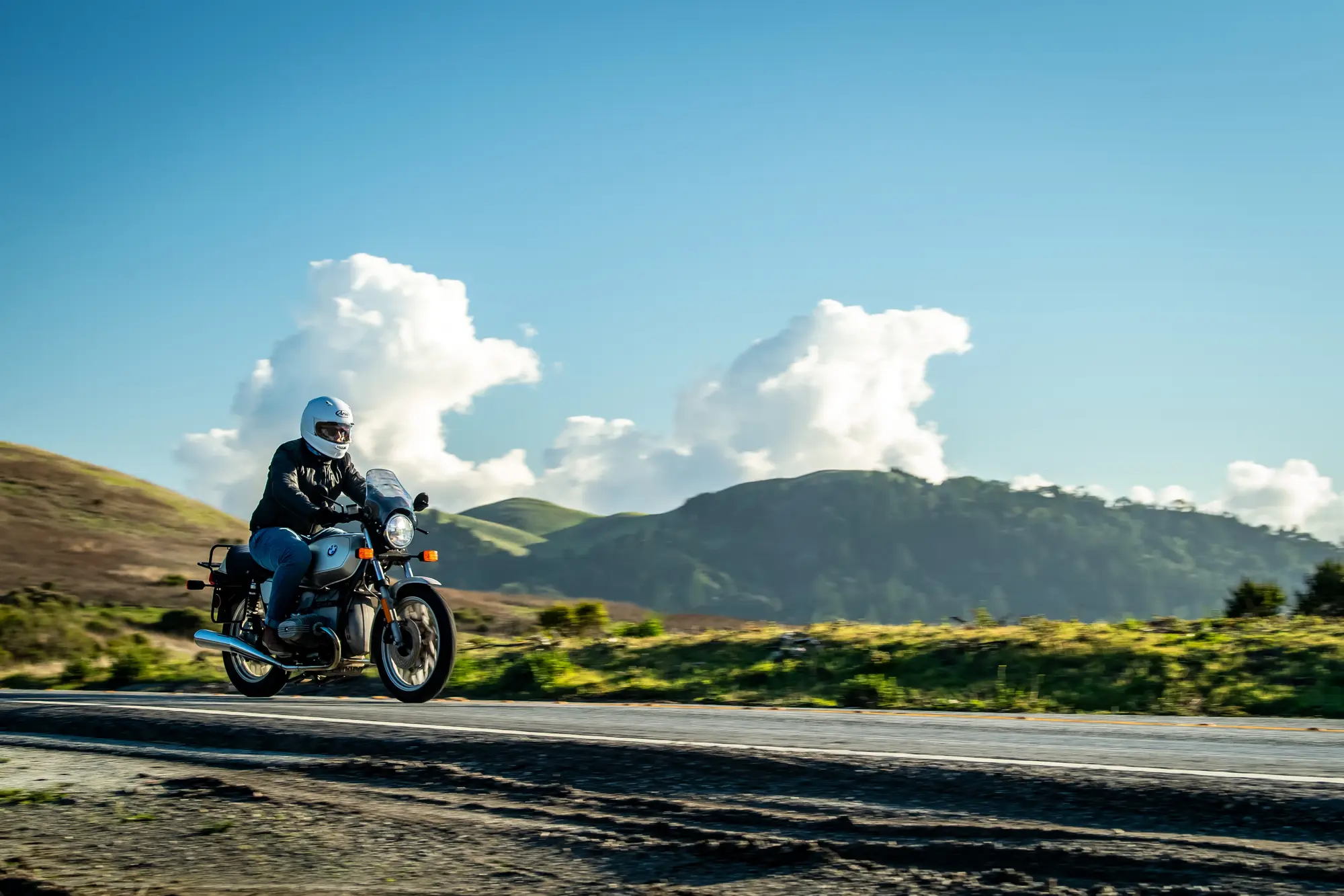 A rider on a classic motorcycle on a country road below green hills