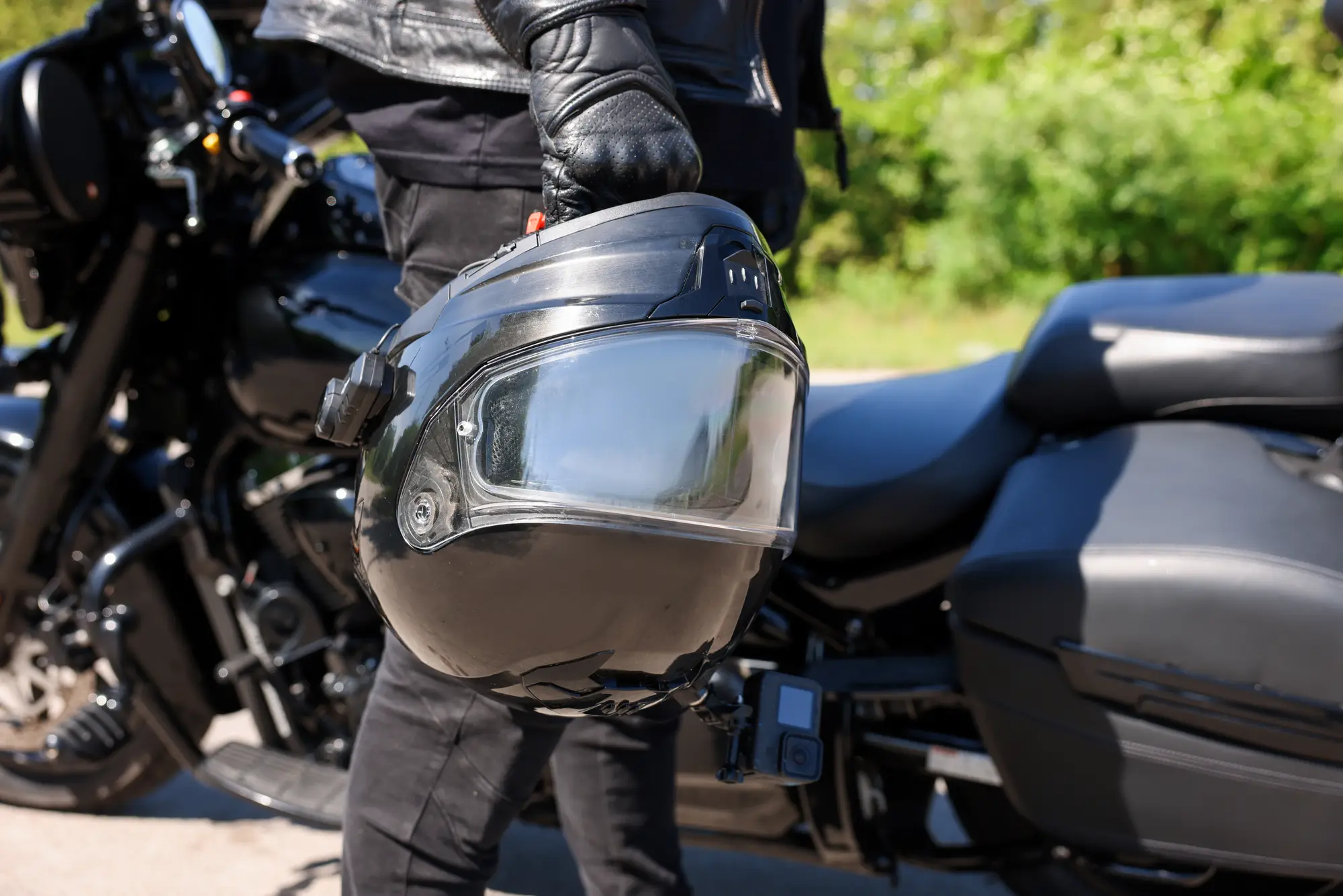 A rider in a leather jacket holding a black full-face helmet beside a motorcycle